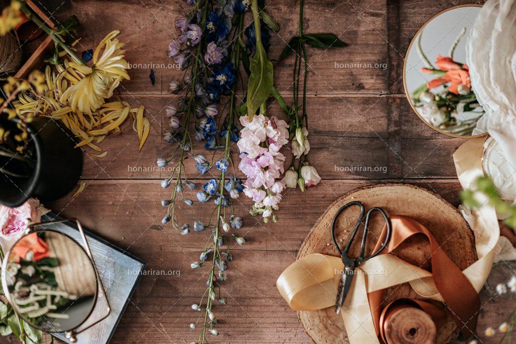 عکس با کیفیت انواع گل بر روی میز چوبی به همراه ربان و قیچی